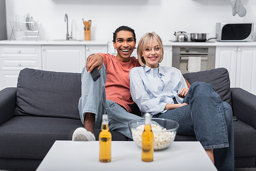 happy interracial couple watching movie near beer bottle and popcorn on coffee table