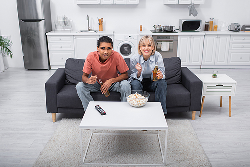 emotional interracial couple cheering and holding bottles with beer while watching sport match in living room