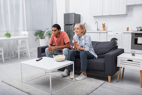 young multiethnic couple holding bottles with beer while watching sport match in living room