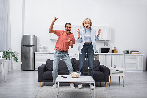 happy multiethnic couple jumping while watching sport match in living room