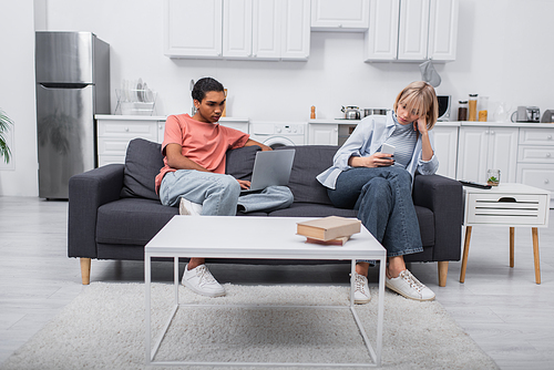 african american freelancer using laptop near girlfriend with smartphone sitting on couch