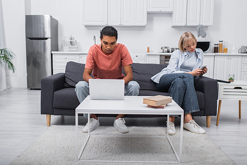 african american man using laptop near girlfriend with smartphone sitting on couch