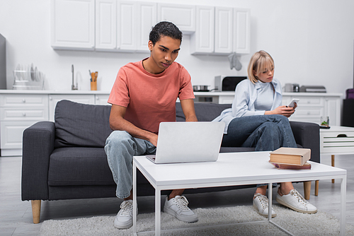 african american man using laptop near blonde girlfriend with cellphone sitting on couch