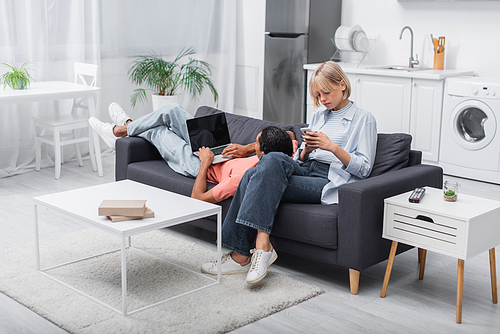 african american man using laptop with blank screen and lying near blonde girlfriend with cellphone on couch