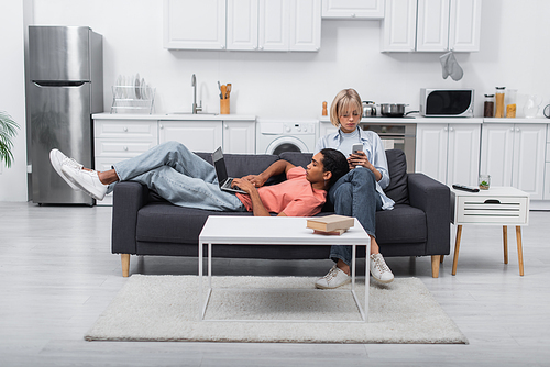 african american man using laptop with blank screen and lying near blonde girlfriend with smartphone on couch