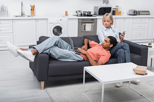 blonde girlfriend holding smartphone and looking at african american man working on laptop