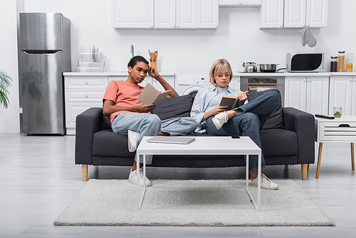 young african american man and blonde woman reading books near gadgets on coffee table