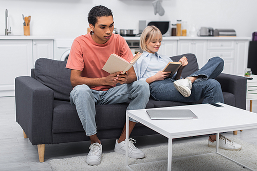 young african american man and blurred woman reading books near gadgets on coffee table