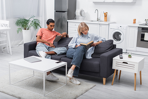 young african american man and blonde woman reading books near gadgets in living room