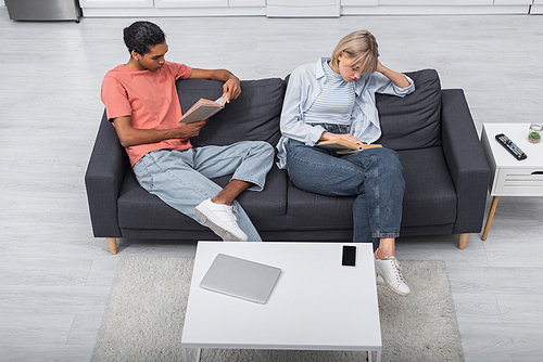 top view of young african american man and blonde woman reading books near gadgets in living room