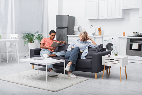 young african american man and blonde woman reading books near gadgets in modern living room