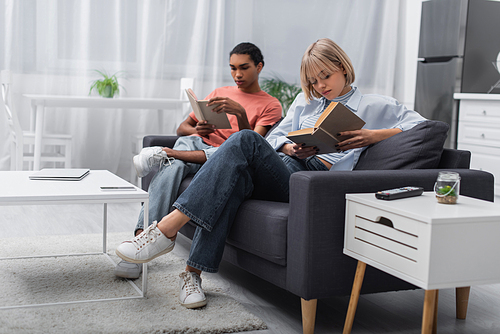 young blonde woman and african american man reading books near gadgets in modern living room