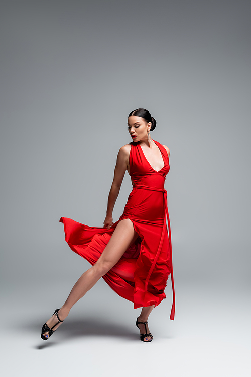 Full length of brunette dancer in red dress looking at leg on grey background