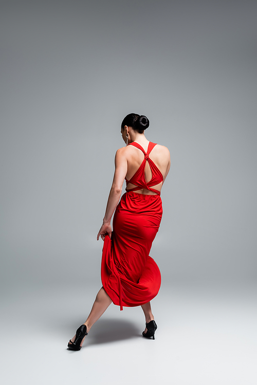 Back view of brunette woman in red dress and heels dancing tango on grey background