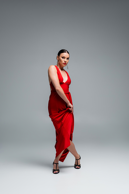 Stylish ballroom dancer in red dress and heels looking at camera on grey background