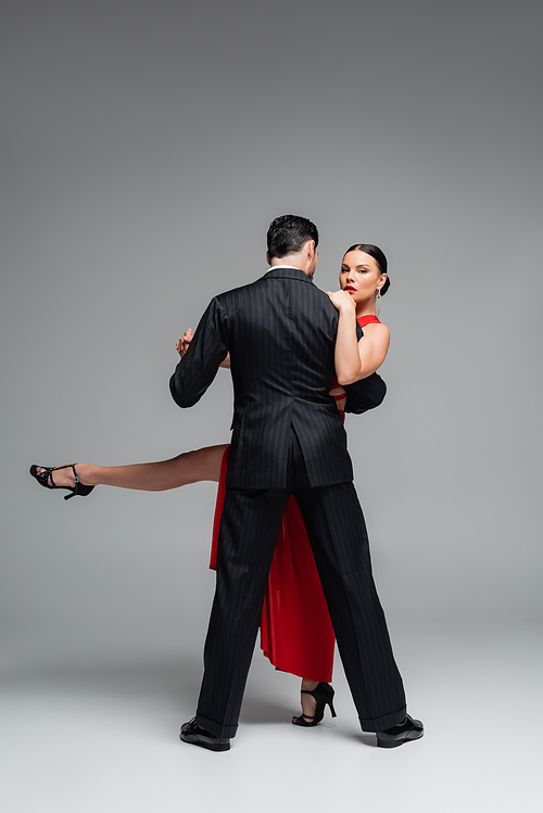 Elegant woman in heels looking at camera while dancing tango with partner on grey background