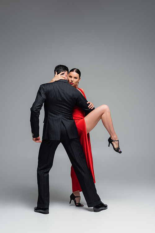 Elegant woman in red dress touching partner while dancing tango on grey background