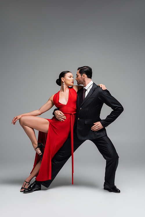 Elegant woman in dress looking at partner while dancing tango on grey background