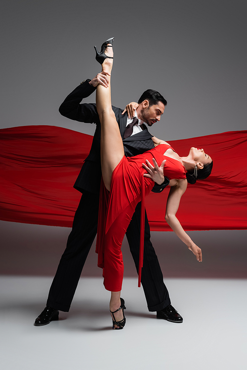 Side view of elegant dancer touching leg of partner while performing tango on grey background with red fabric