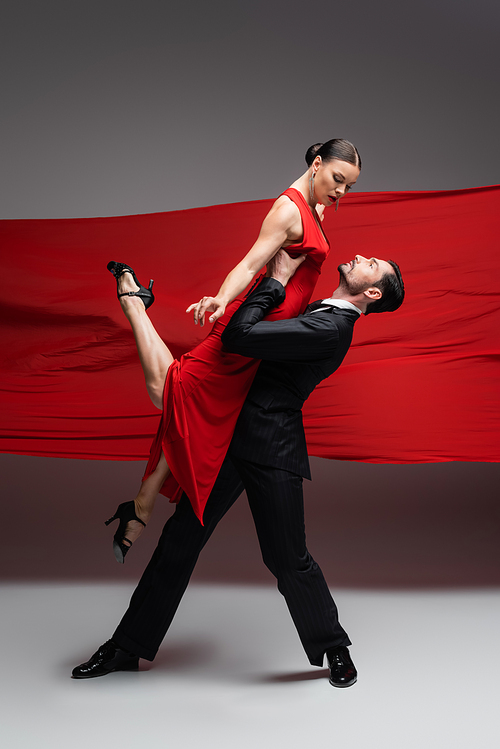 Dancer in suit lifting elegant partner while performing tango on grey background with red fabric