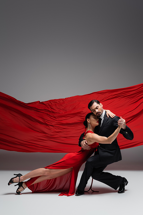 Elegant couple holding hands while performing tango on grey background with red fabric