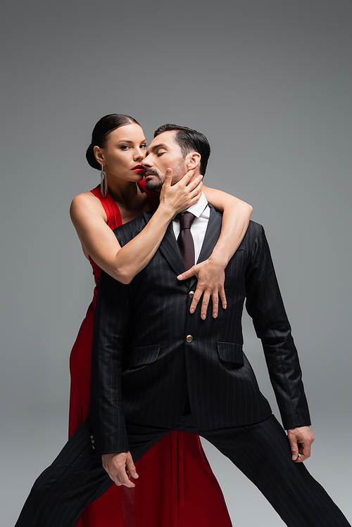 Stylish woman in red dress touching man in suit and looking at camera isolated on grey