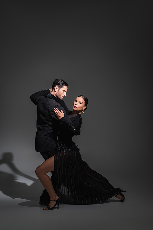 Elegant woman with red lips looking at camera while dancing tango with partner on grey background with shadow