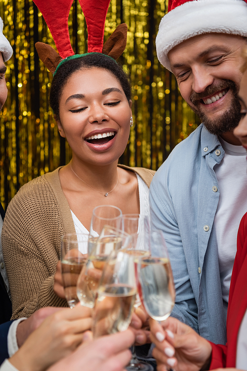 Cheerful multiracial businesswoman in Christmas headband clinking champagne with colleagues in office