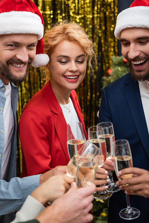 Businesswoman clinking champagne with smiling interracial colleagues in santa hats in office