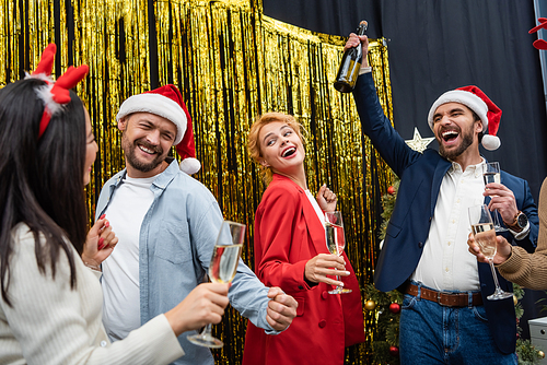 Excited businessman in santa hat holding bottle of champagne near interracial colleagues dancing in office