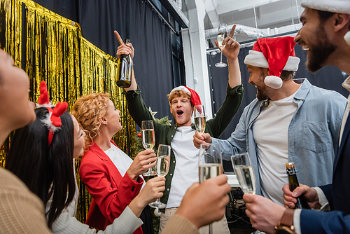 Excited businessman in santa hat holding champagne while celebrating Christmas with multiethnic colleagues in office