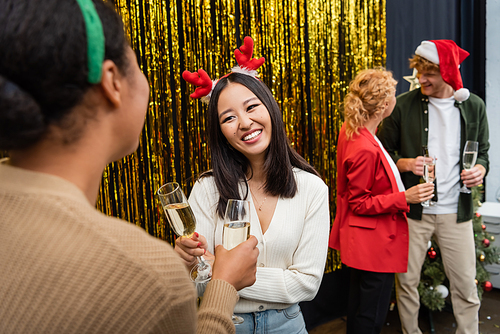 Smiling asian businesswoman holding champagne and talking to multiracial colleague during Christmas party in office
