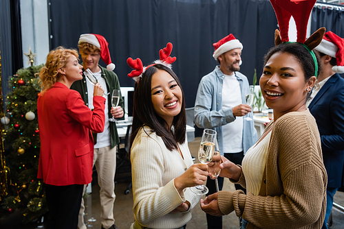Smiling multiethnic businesswomen in Christmas headbands holding champagne and looking at camera in office