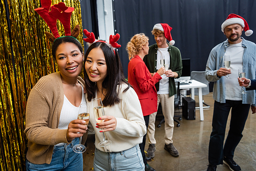 Positive interracial businesswomen in Christmas headbands holding champagne near tinsel in office