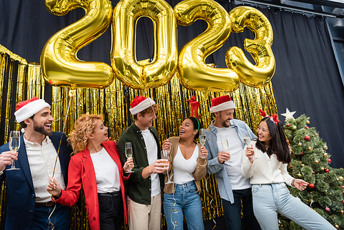 Smiling multiethnic business people holding champagne and balloons in shape of 2023 signs in office