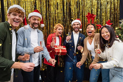 Excited multiethnic business people holding champagne and sparklers under confetti during Christmas party in office