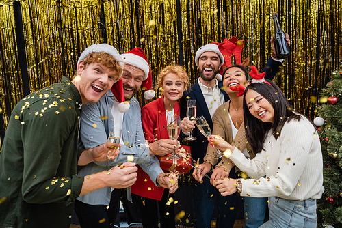 Smiling multiethnic business people in santa hats holding champagne and sparklers under confetti in office