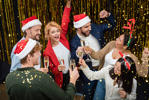 Positive interracial business people holding champagne and dancing under confetti during Christmas party in office