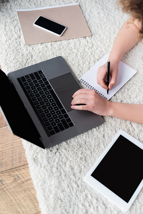 top view of cropped woman writing in notebook near devices with blank screen on floor
