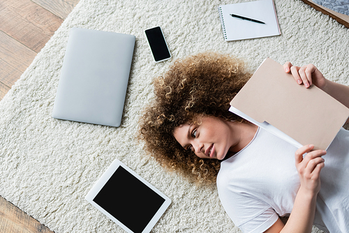 top view of curly woman with folder lying on floor carpet near devices and notebook
