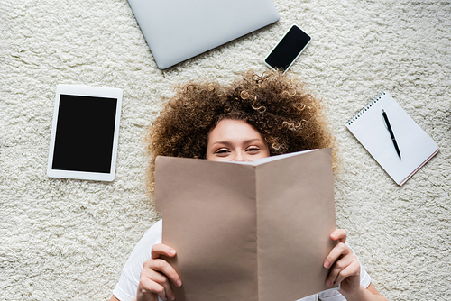top view of cheerful curly woman obscure face with folder near gadgets and notebook