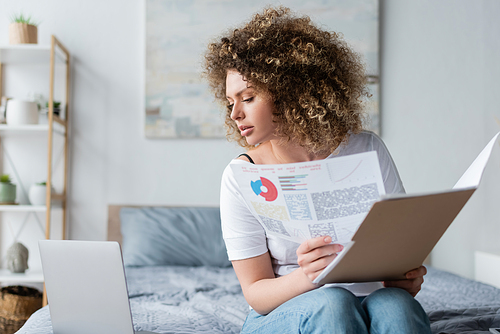 curly woman with charts and folders looking at laptop on bed at home