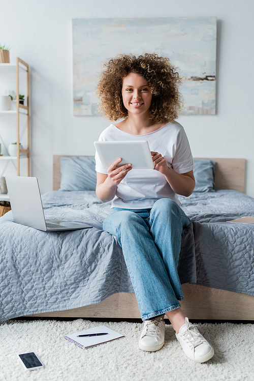 happy woman with digital tablet smiling at camera near laptop on bed