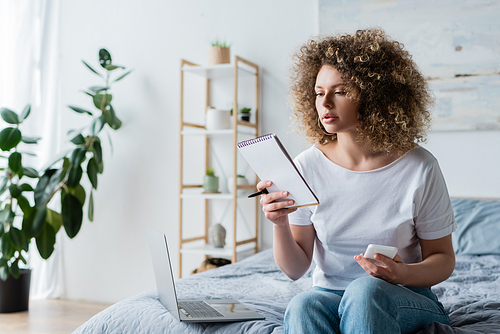 curly woman with smartphone and notebook sitting on bed near laptop