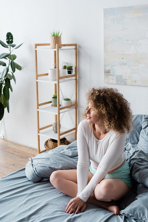 curly woman sitting on bed with crossed legs near blurred rack with flowerpots