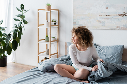 positive woman in white pajamas sitting on grey bedding near rack with flowerpots