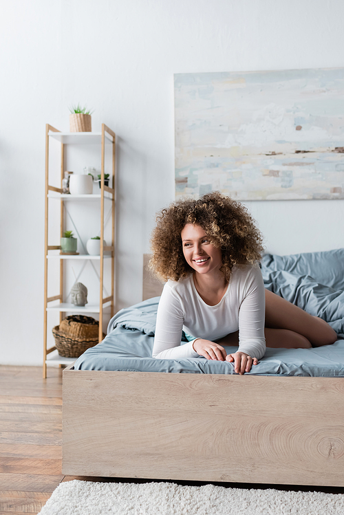 young curly woman smiling on bed near blurred rack with potted plants