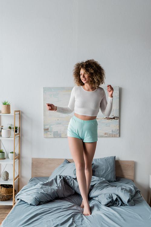 full length of happy young woman in white pajamas standing on bed at home