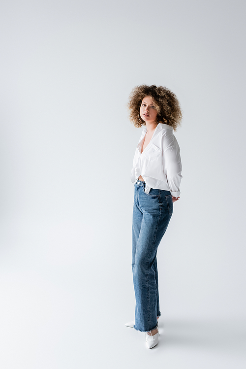 Curly woman in blouse and jeans looking at camera on white background