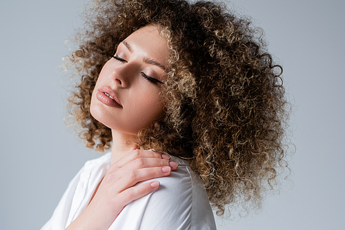 Young curly model touching shoulder and closing eyes isolated on grey